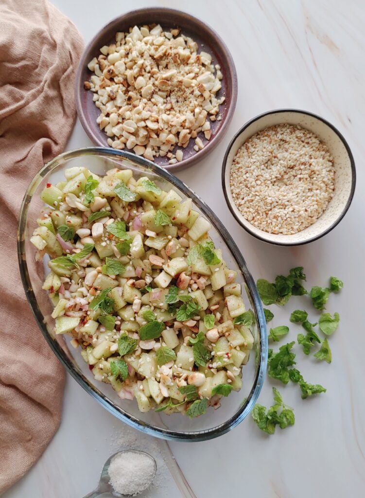 cucumber salad served in an oval-shaped bowl kept on the table with crushed peanuts, sesame seeds, mint leaves, and salt on the side